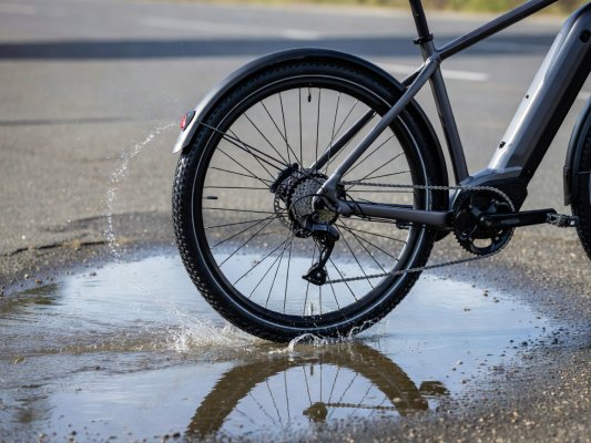 Professional realistic photograph, natural daylight: Close up of a genuine full length mudguard on a real bicycle wheel riding through a realistic puddle. Accurate wheel spokes, natural water spray, no AI artifacts.
